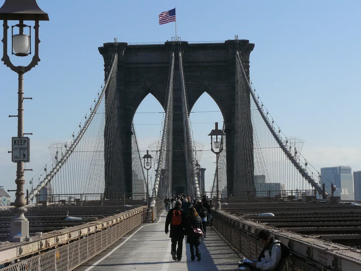 Pedestrians on the wooden promenade of the Brooklyn Bridge below its Gothic stone towers, on a New York architecture tour